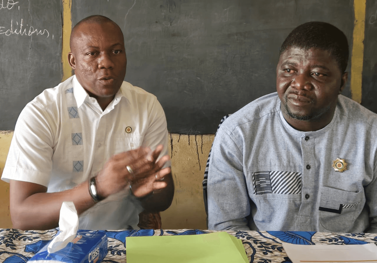 L'Honorable Innocent Yao et un intervenant assis à la table de réunion devant un tableau d'école.