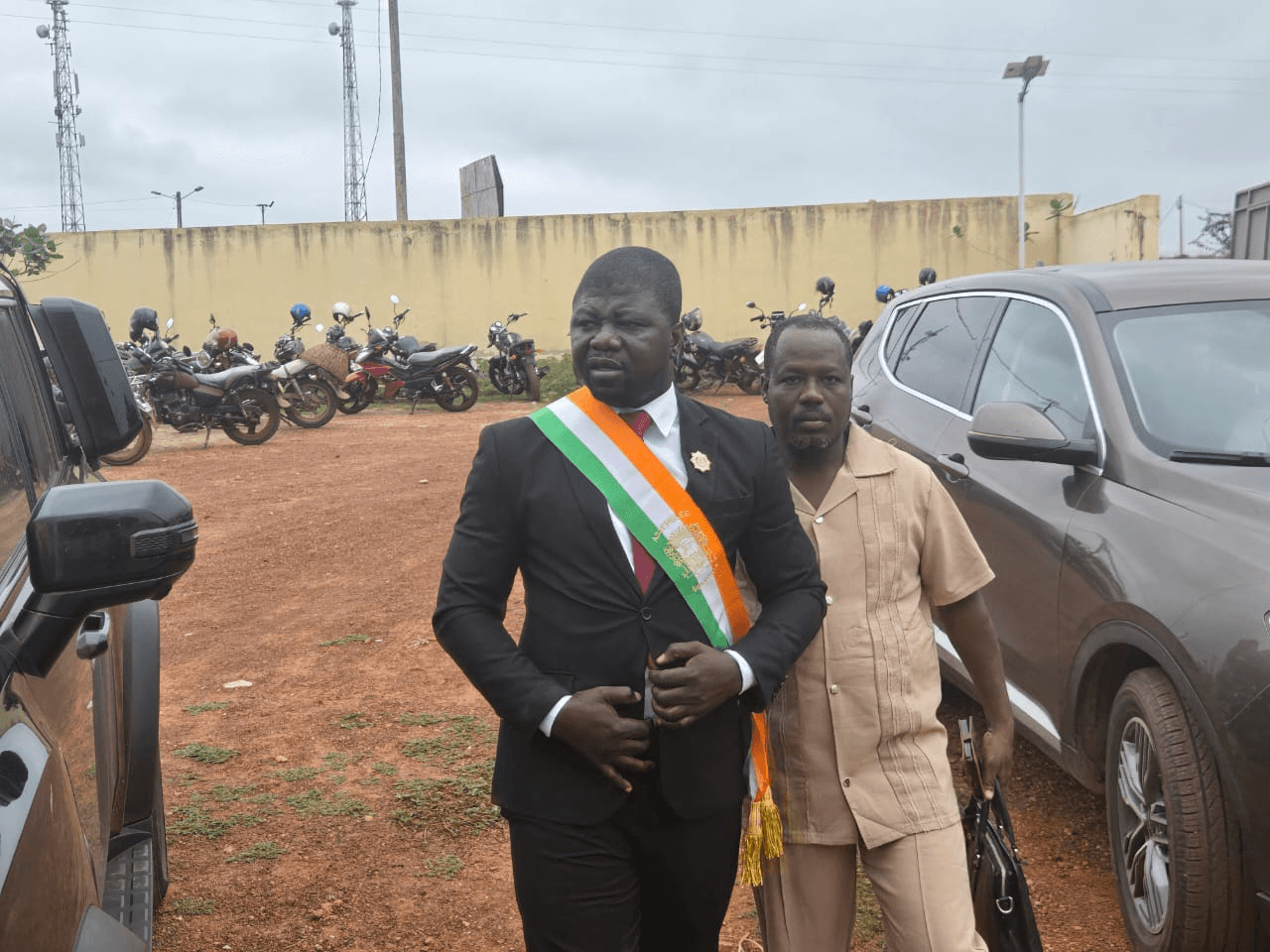 L'Honorable Innocent Yao en costume et écharpe tricolore avec un collaborateur en extérieur.