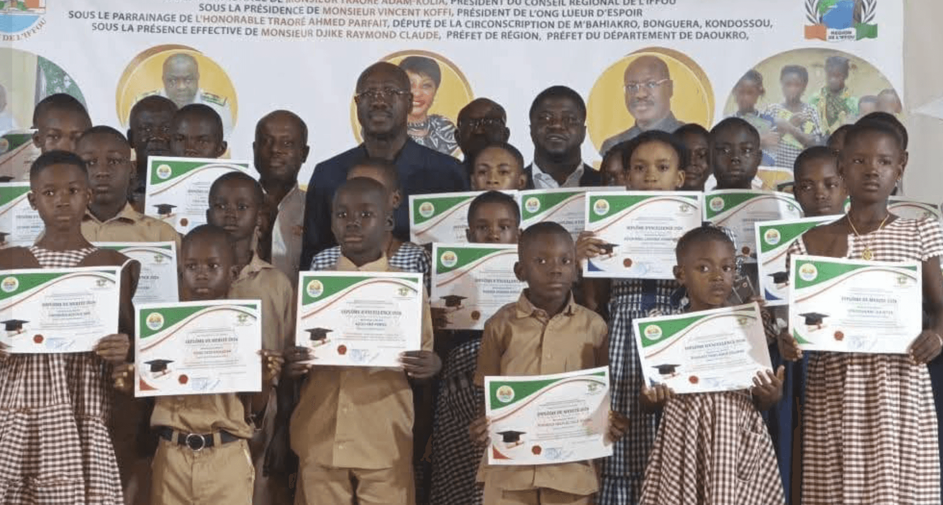 Élèves en uniforme scolaire présentant leurs attestations lors d'une cérémonie de remise de prix, adultes et banderole en arrière-plan.