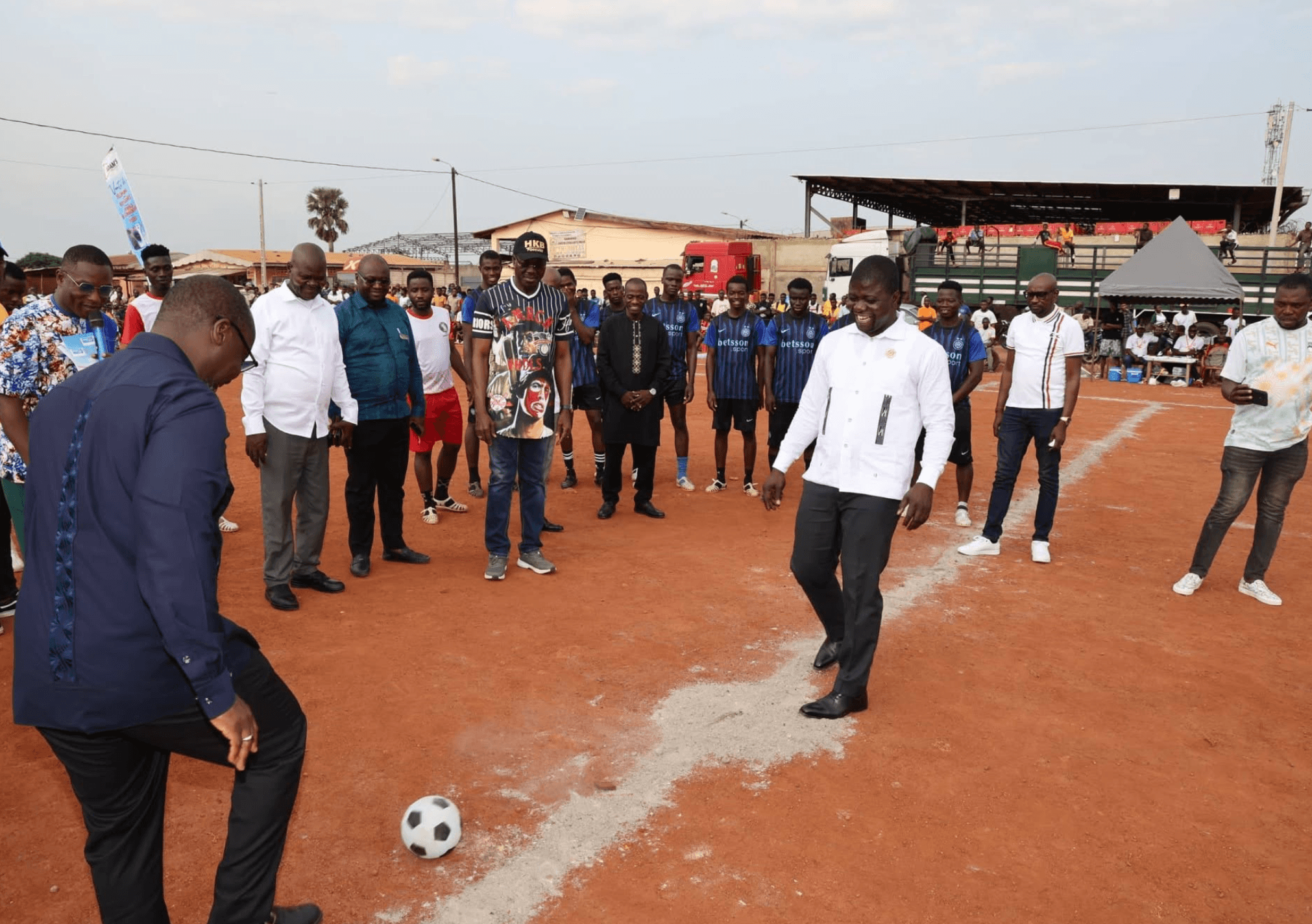 Coup d'envoi d'un match de football sur un terrain communautaire, en présence d'invités officiels.